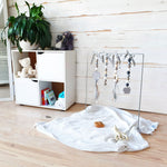 A collapsible white mobile frame made of steel with rubber coating, designed for infant play. Toys attached to frame in a play setting for tummy time play.