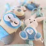 A close up of a set of four handcrafted felt dingle dangle toys in woodland animal shapes in shades of blue: a rabbit, an owl, a fox, and a raccoon. Made from soft felt with ribbons, designed for infant sensory development.