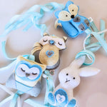 A birds eye view of a set of four handcrafted felt dingle dangle toys in woodland animal shapes in shades of blue: a rabbit, an owl, a fox, and a raccoon. Made from soft felt with ribbons, designed for infant sensory development.